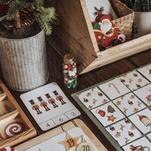 Load image into Gallery viewer, A Christmas morning basket setup with various holiday-themed items including cards, a memory game, and decorative elements on a wooden table.